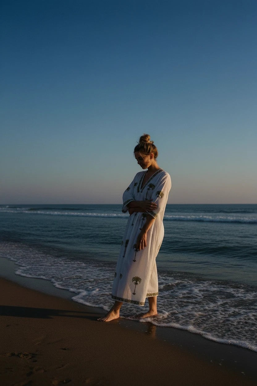 Woman wearing The House of Saffron white midi dress with olive-green palm tree embroidery on a beach at sunset.