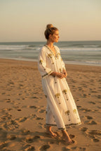 The House of Saffron white midi dress with olive green palm tree embroidery, V-neckline, and patterned trim on beach.