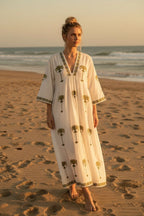 The House of Saffron white midi dress with olive green palm tree embroidery, V-neckline, and flowy fit on a beach.